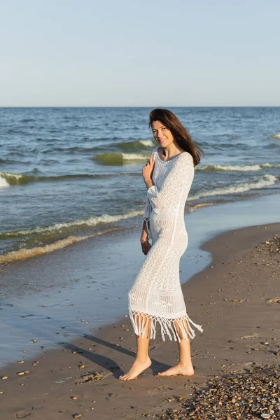 Barefoot woman in knitted dress standing on beach near sea