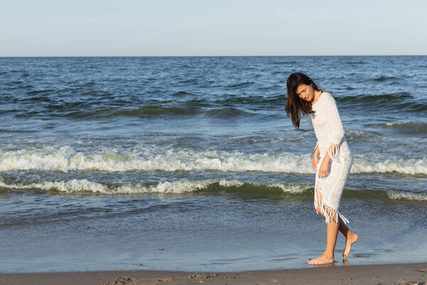 Full length of brunette woman in dress walking on sand near sea 