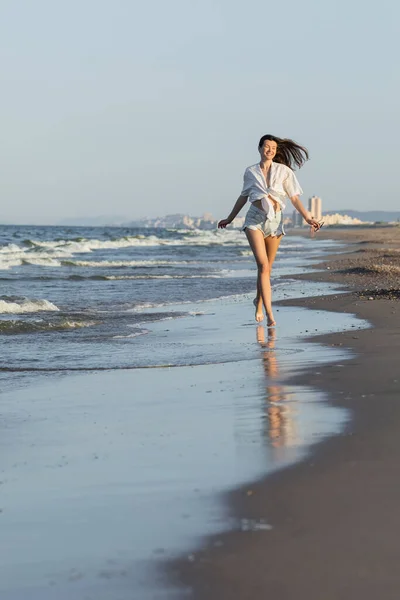 Cheerful brunette woman in shorts holding sunglasses while running on beach