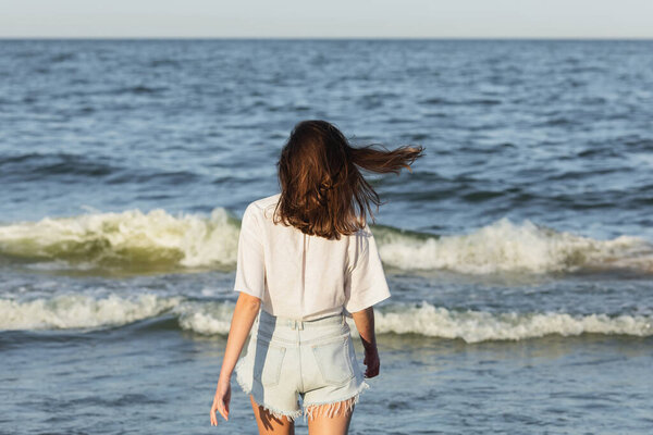 Back view of brunette woman in denim shorts standing near blurred sea 