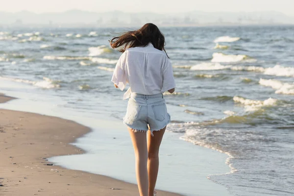 Back view of woman in white shirt and denim shorts walking near sea on beach