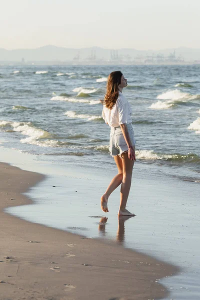 海の近くの湿った砂の上に立つショートパンツの若い女性のサイドビュー — ストック写真