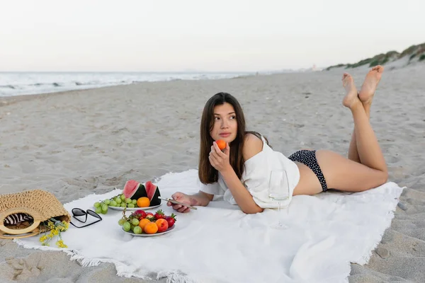 Μελαχρινή Γυναίκα Μαγιό Και Πουκάμισο Κρατώντας Smartphone Και Φρούτα Κοντά — Φωτογραφία Αρχείου