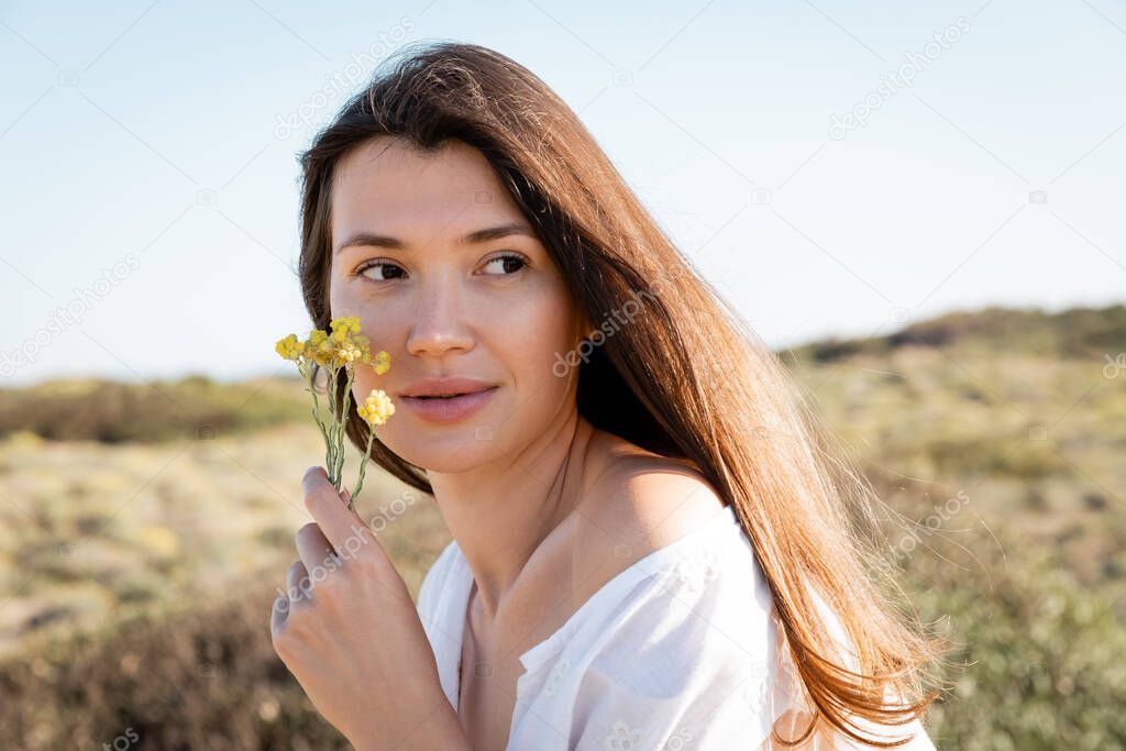Brunette woman holding yellow flowers near face on blurred beach 