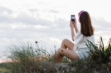 Kulaklıklı bir kadının akşamleyin sahilde akıllı telefonuyla fotoğraf çekişini. 