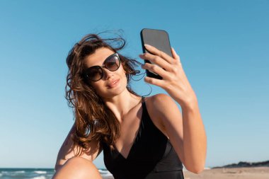 Mayolu ve güneş gözlüklü genç bir kadın sahilde akıllı telefondan selfie çekiyor.