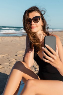 Mayolu ve güneş gözlüklü genç bir kadın deniz kenarında cep telefonundan selfie çekiyor.