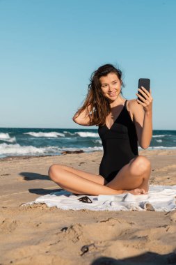Mayolu mutlu genç bir kadın sahilde otururken telefonuyla selfie çekiyor.