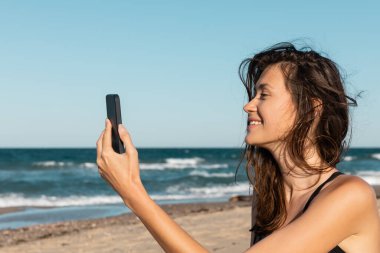 Mayo giymiş neşeli genç bir kadın deniz kenarındaki akıllı telefondan selfie çekiyor.