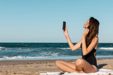 Deniz kenarında battaniyede otururken mayo giymiş esmer bir kadın selfie çekiyor.