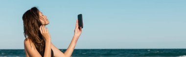 Bikini giymiş esmer bir kadın deniz kenarındaki akıllı telefondan selfie çekiyor.