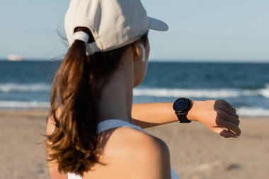 Beyzbol şapkalı esmer kadının fitness takip cihazını kontrol edişinin arka görüntüsü 