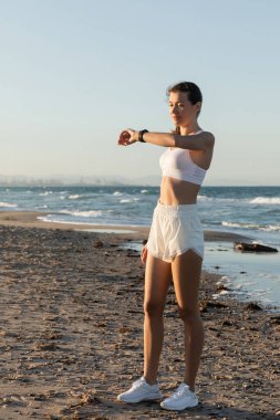 Beyaz spor sutyen ve şortlu genç bir kadın sahilde deniz kenarında spor takip cihazına bakıyor.