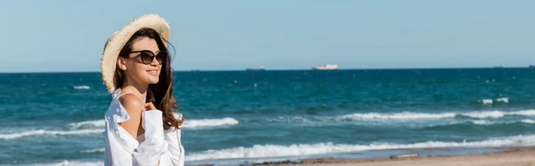 Güneş gözlüklü, hasır şapkalı, deniz kenarında beyaz gömlekli gülümseyen kadın.