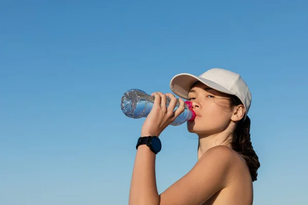 Спортивная Женщина Бейсболке Пьет Освежающую Воду Против Голубого Неба — стоковое фото