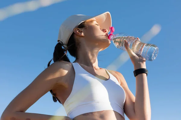 野球帽のスポーツ女性と青空に対するスポーツブラジャー飲料水の低角度ビュー — ストック写真