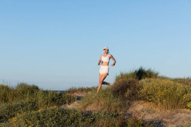 young sportive woman in baseball cap and wireless earphones running on beach clipart