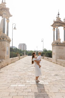 Side view of woman in white dress hugging toddler daughter on Puente Del Mar bridge in Valencia