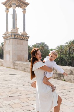 Excited mom embracing baby girl in summer dress on Puente Del Mar bridge in Valencia