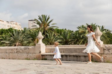 Side view of mom and daughter in summer dresses running on Puente Del Mar bridge in Valencia clipart