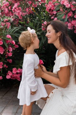 Smiling woman holding flower and touching baby daughter in summer dress in Valencia