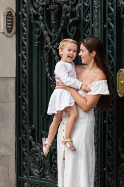 Smiling woman holding baby daughter near building on street in Valencia
