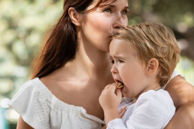 Woman kissing daughter with cookie in summer park 