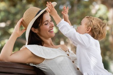 Toddler girl playing with mom in summer hat on bench in park 