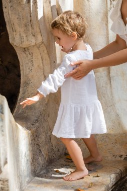 Side view of woman touching toddler daughter in summer dress on stone bench in Valencia