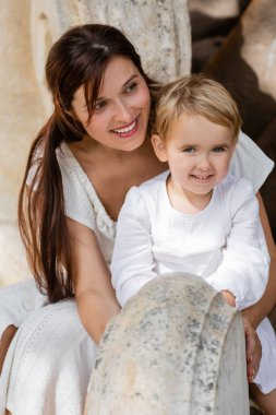 Beyaz elbiseli gülümseyen anne ve çocuk Valencia 'da parktaki taş bankta oturuyor. 