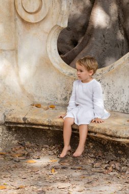 Barefoot baby girl in dress sitting on stone bench in park in Valencia 