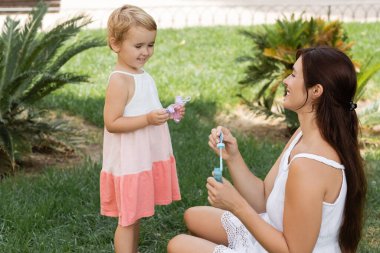 Smiling child holding soap bubbles near mom on green lawn 