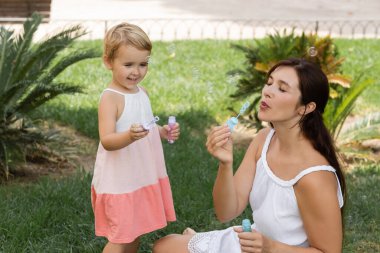 Positive kid holding soap bubbles near mother on lawn 