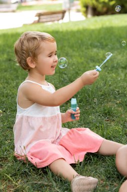 Yazlık elbiseli neşeli çocuk parkta sabun köpüğüyle oynuyor. 
