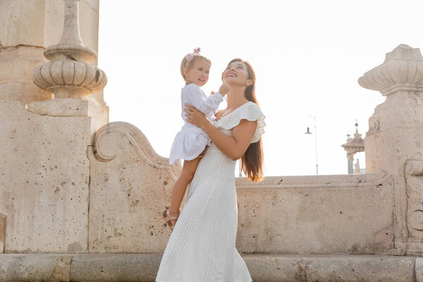 Positive woman in summer dress holding toddler daughter near stone Puente Del Mar bridge in Valencia