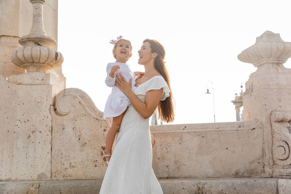 Happy mother holding child in dress near stone Puente Del Mar bridge in Valencia