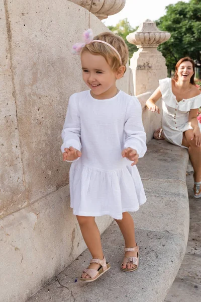 Smiling child in dress walking on stone bench of Puente Del Mar bridge near blurred mom in Valencia