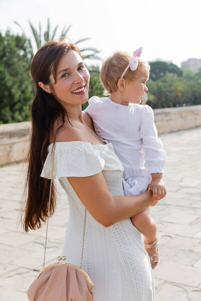 Positive mother in dress looking at camera while holding child on Puente Del Mar bridge in Valencia
