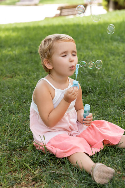 Portrait of toddler kid in dress blowing soap bubbles on lawn 