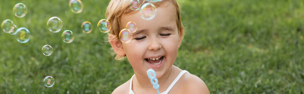 Cheerful baby girl holding wand near soap bubbles in park, banner 