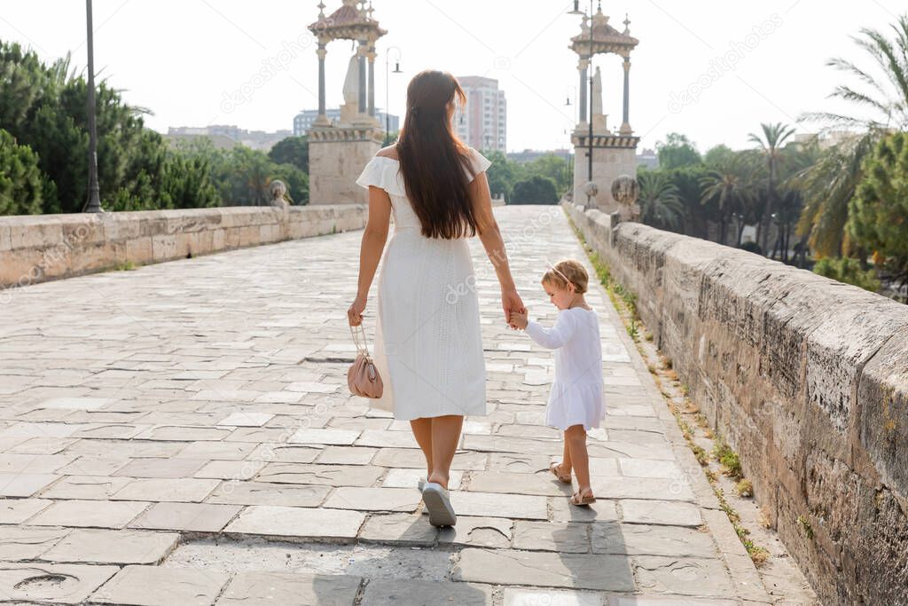 Woman in dress holding hand of toddler daughter while walking on Puente Del Mar bridge in Valencia