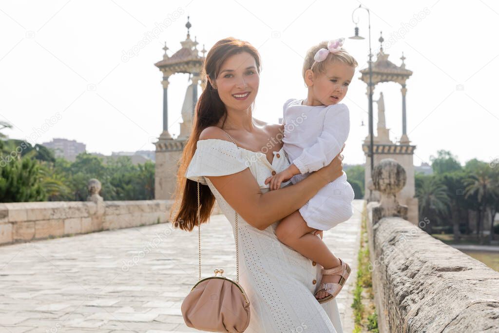 Positive woman holding daughter in dress on Puente Del Mar bridge in Valencia