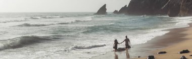 back view of man holding hands with girlfriend in dress while walking on wet sand near ocean water, banner clipart