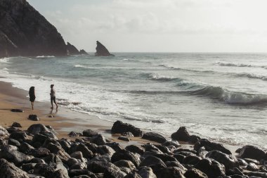 bearded man looking at girlfriend in dress running on wet sand near ocean water  clipart