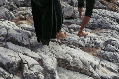 Ayak bileğinde bileklik olan çıplak ayaklı bir adam ve bir kadın kayalıklarda yürüyor.
