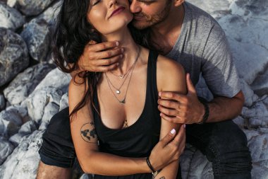 Sakallı bir adamın sahilde kayaların yanında dövmeli bir kadını öptüğü görüntüsü.