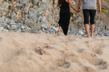 cropped view of couple holding hands and walking on sand near rocks  clipart
