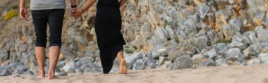 cropped view of couple holding hands and walking on sand near rocks, banner clipart