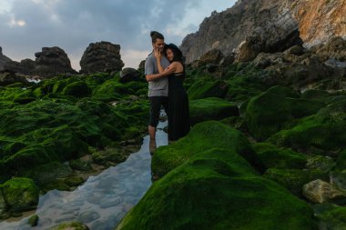 Şapkalı ve elbiseli bir kadın okyanustaki yosunlu taşların yanında sakallı bir adama sarılıyor.