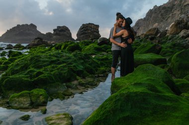 tattooed woman in hat and dress hugging bearded boyfriend near mossy stones in ocean clipart